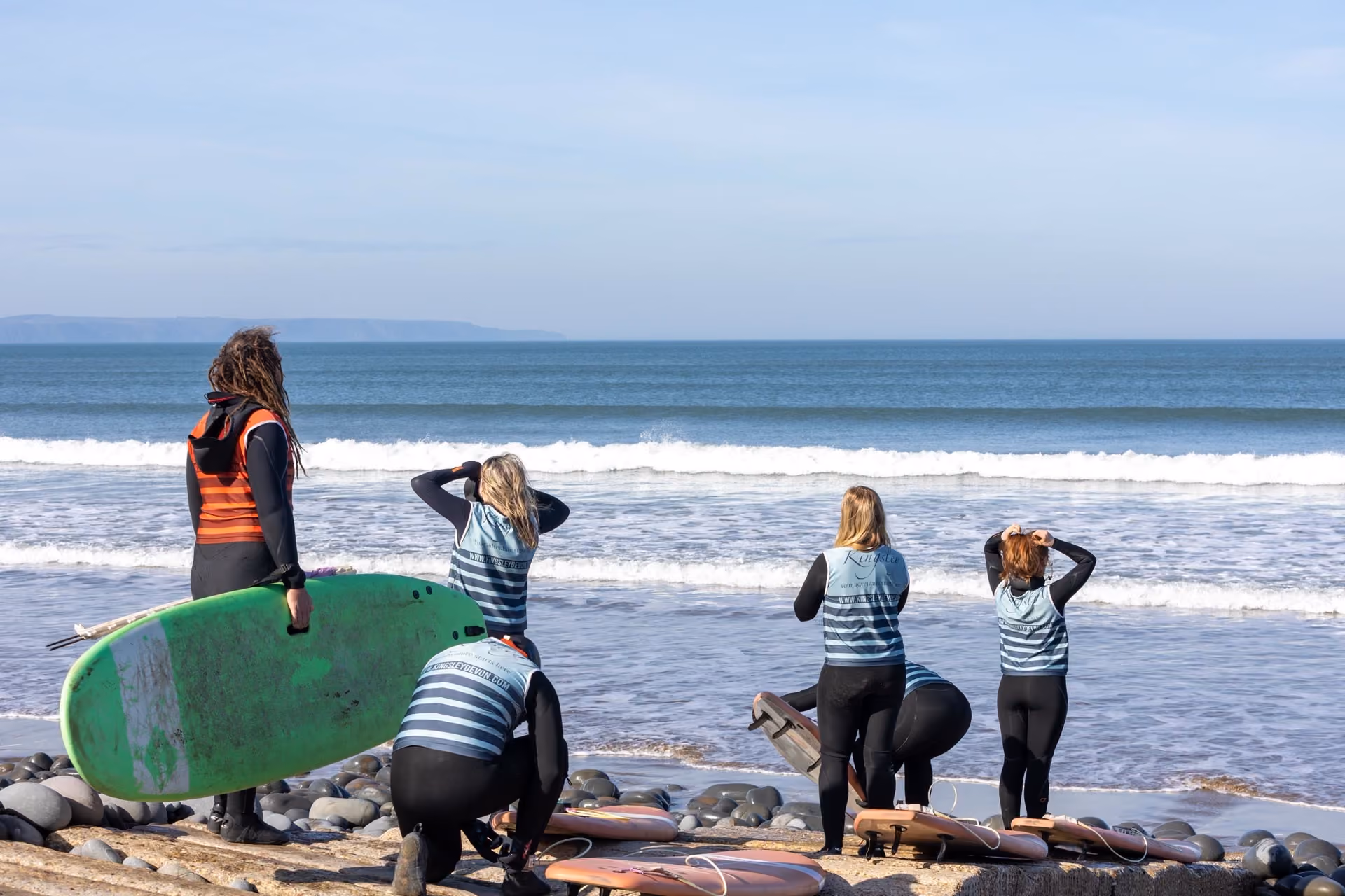 A group of five people wearing wetsuits and striped vests stand on a rocky beach facing the ocean, one holding a green surfboard, preparing to surf.