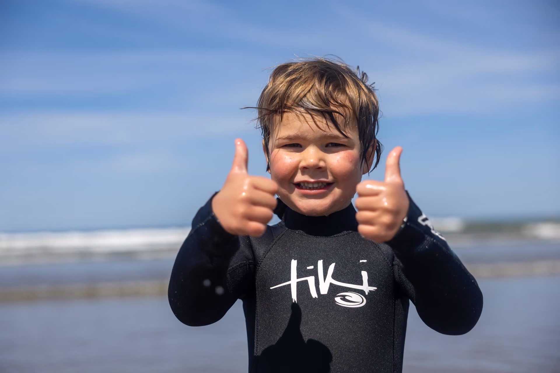 Smiling young child with wet hair wearing a black wetsuit giving two thumbs up, standing on a beach with waves and blue sky in the background.