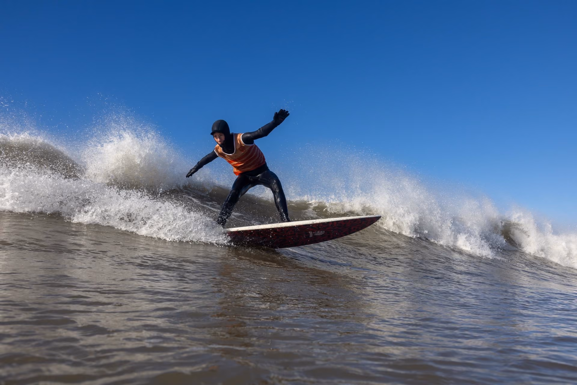 Surfer wearing a black wetsuit and orange vest riding a wave on a surfboard under clear blue sky.
