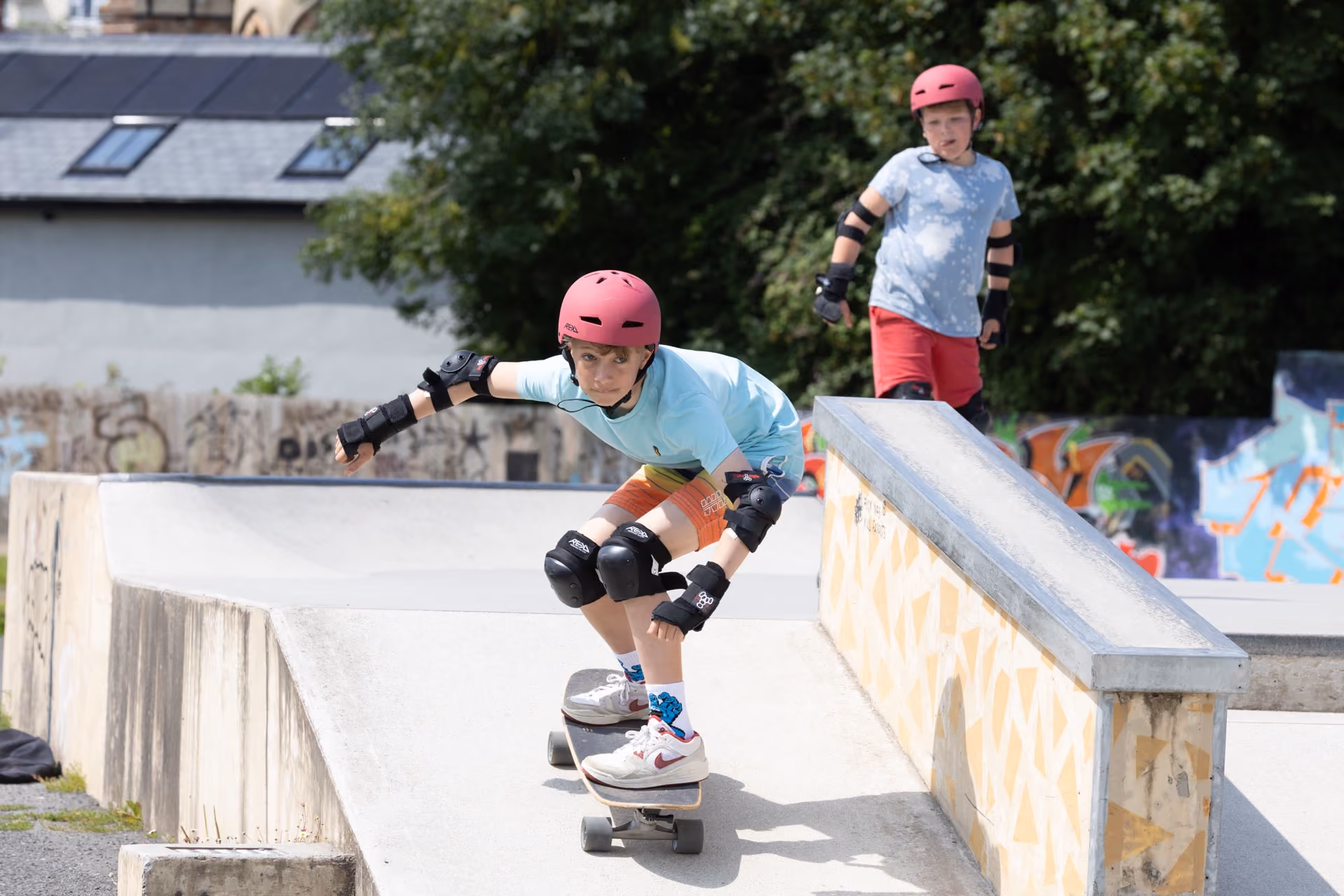 A boy wearing a red helmet and protective pads skateboarding down a concrete ramp in a skatepark, with another boy in a red helmet and blue shirt watching in the background.