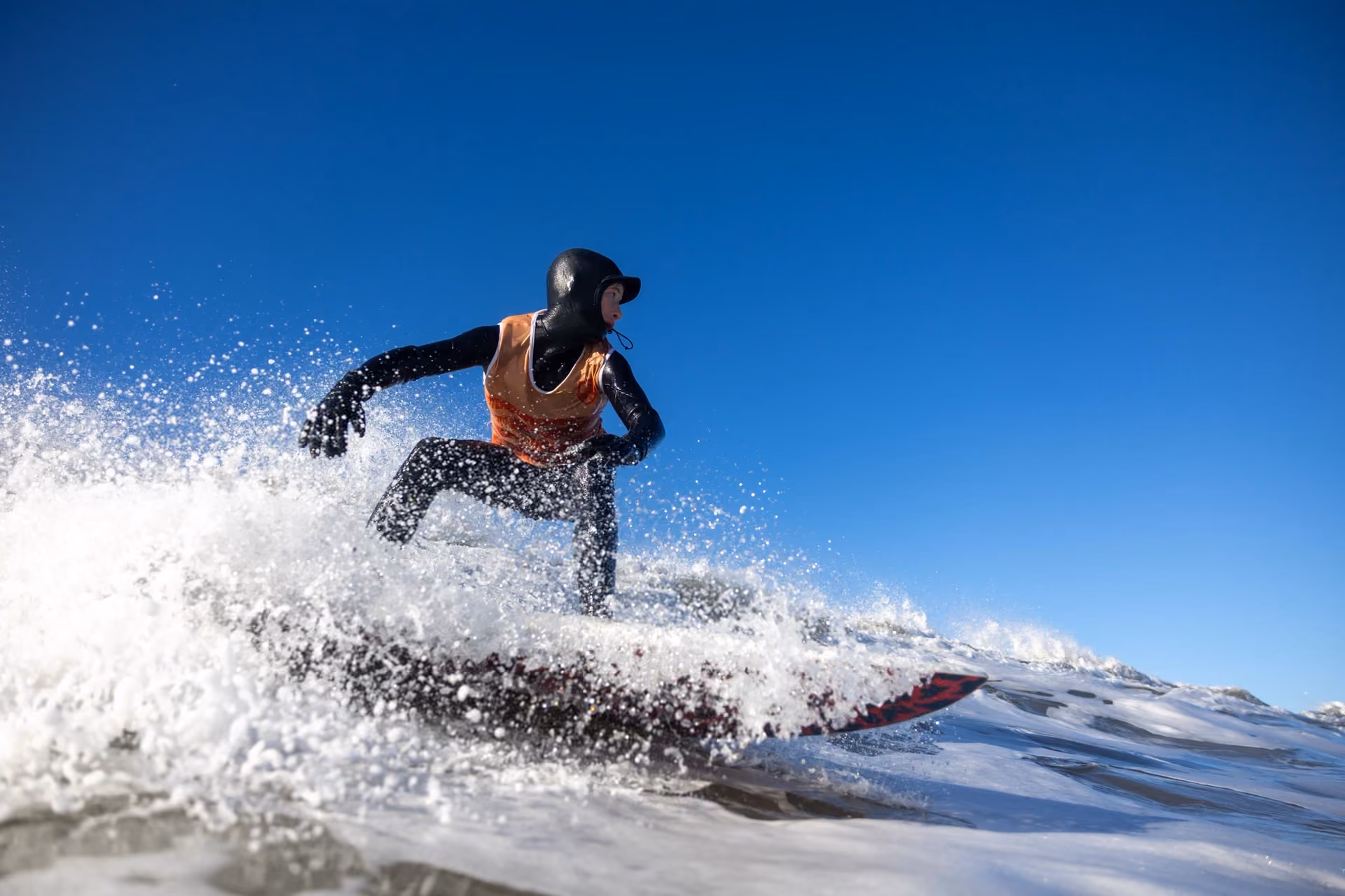 Surfer wearing a black wetsuit and orange vest riding a wave on a surfboard against a clear blue sky.