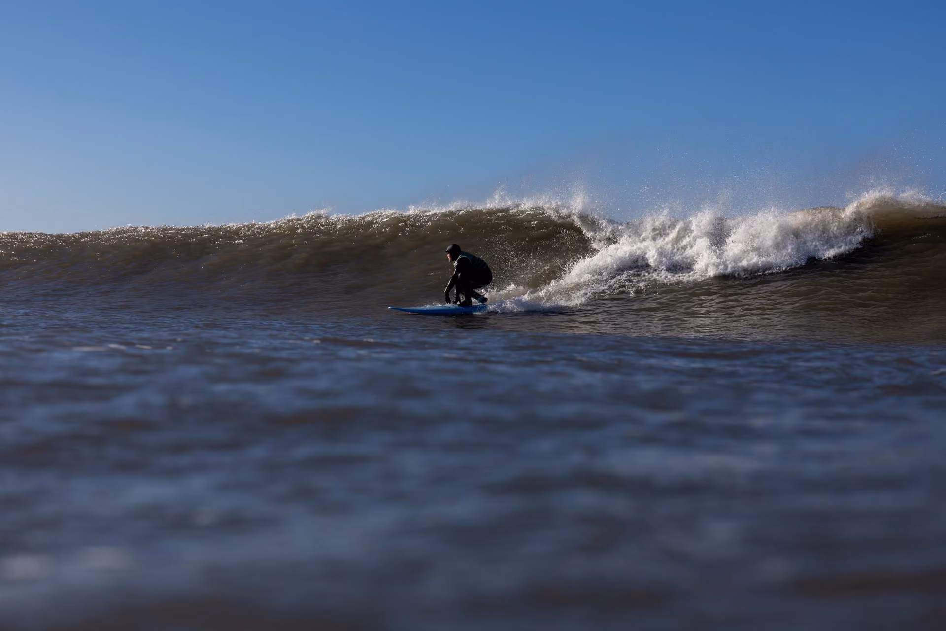 Surfer in a wetsuit riding a large wave on a blue surfboard under a clear blue sky.