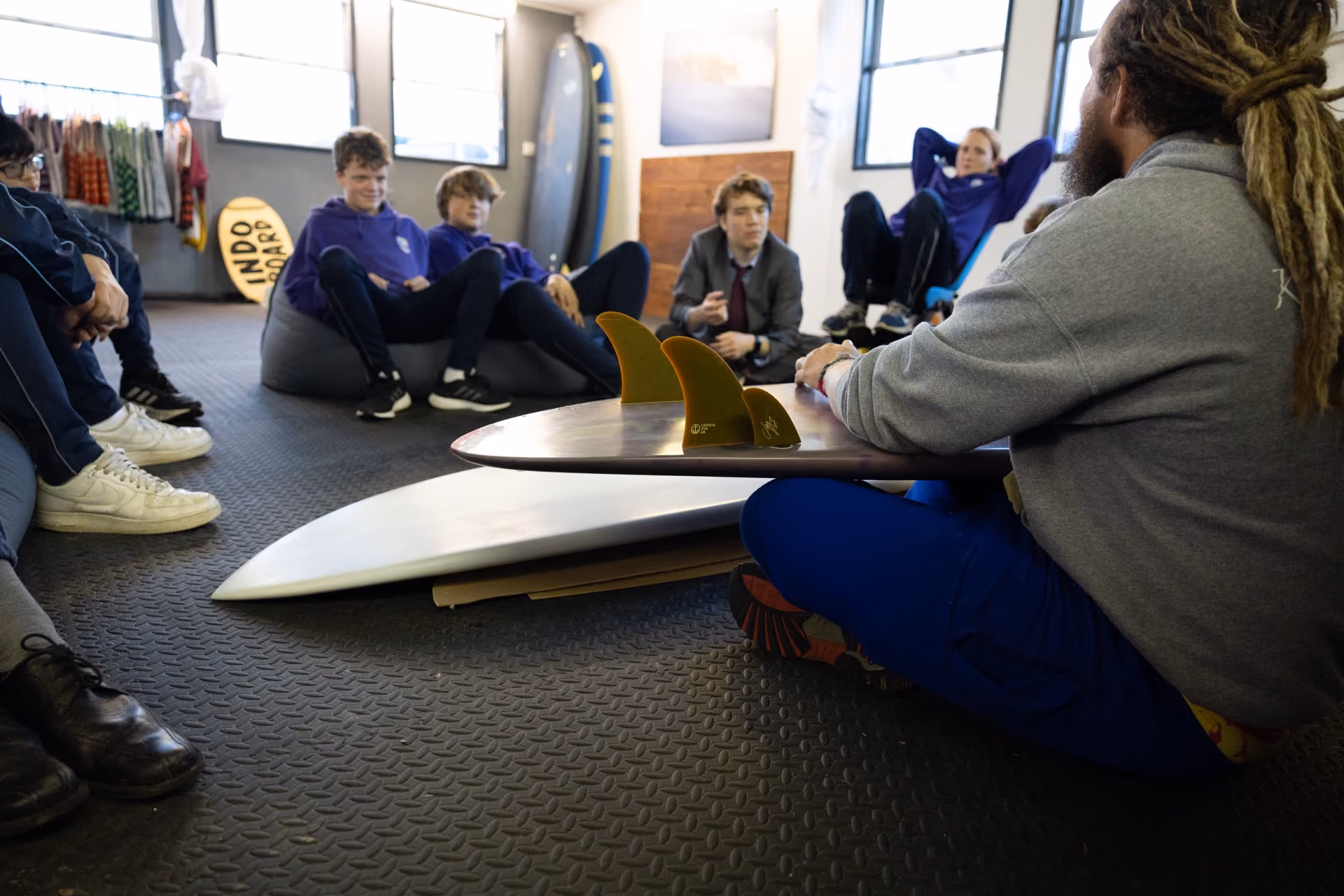 Group of young people sitting on the floor and bean bags in a room, listening to a person with dreadlocks holding a surfboard with visible fins.