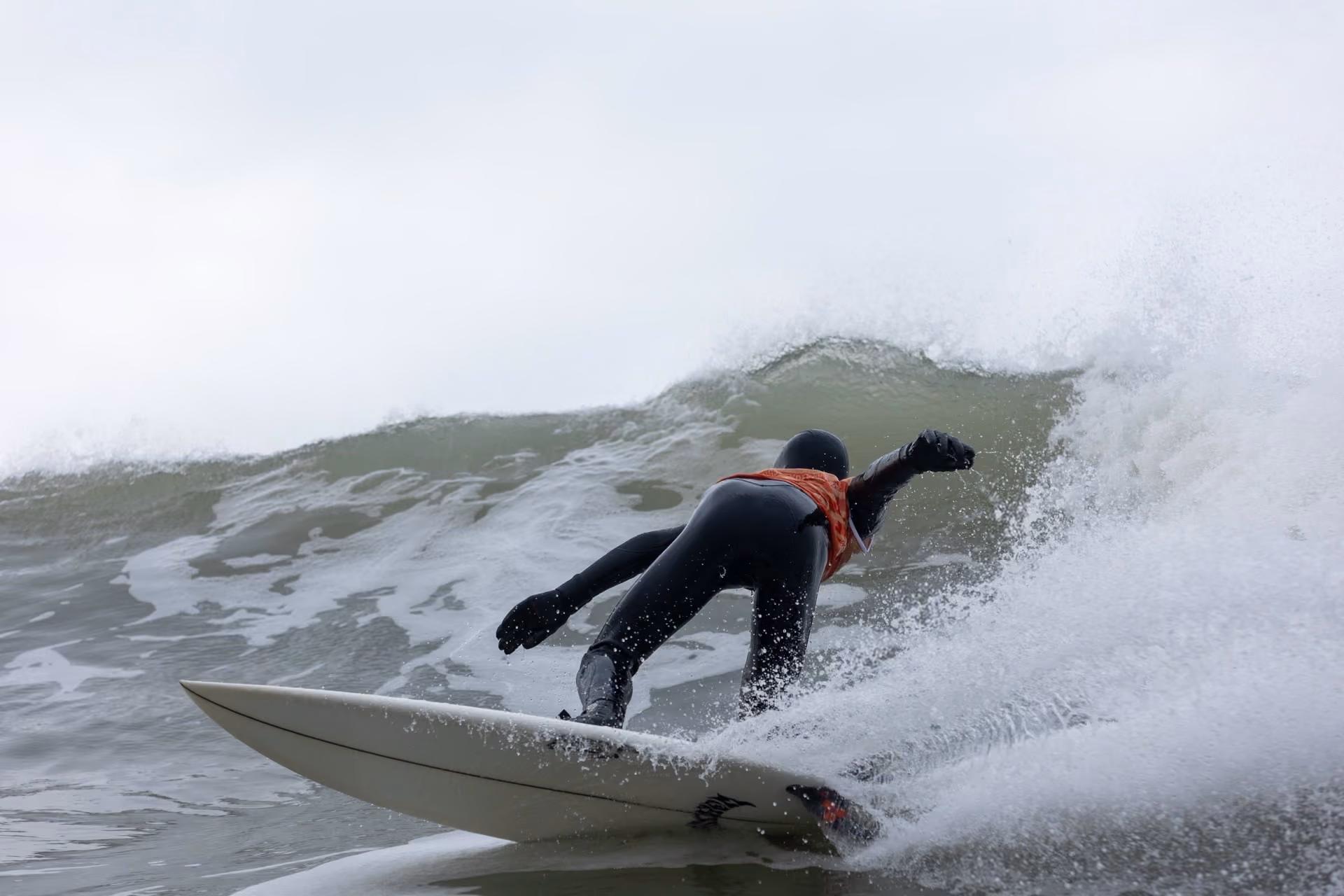 Surfer in a black wetsuit and helmet rides a large wave on a white surfboard, leaning forward with one arm outstretched.
