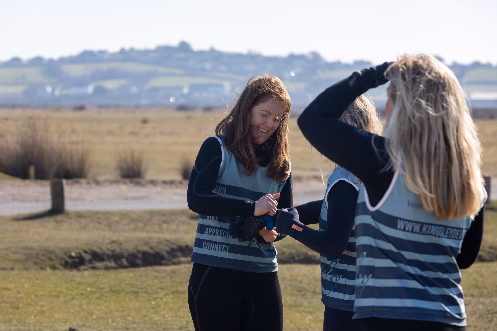 Three women outdoors in a field on a sunny day, wearing matching sleeveless sports tops over long-sleeve shirts, smiling and interacting with each other.