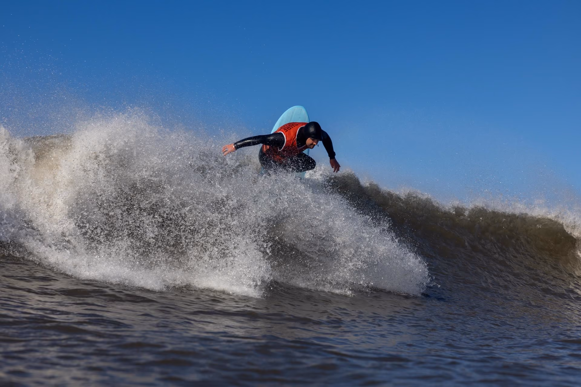 Surfer wearing a black wetsuit and red vest riding a wave with white spray under a clear blue sky.