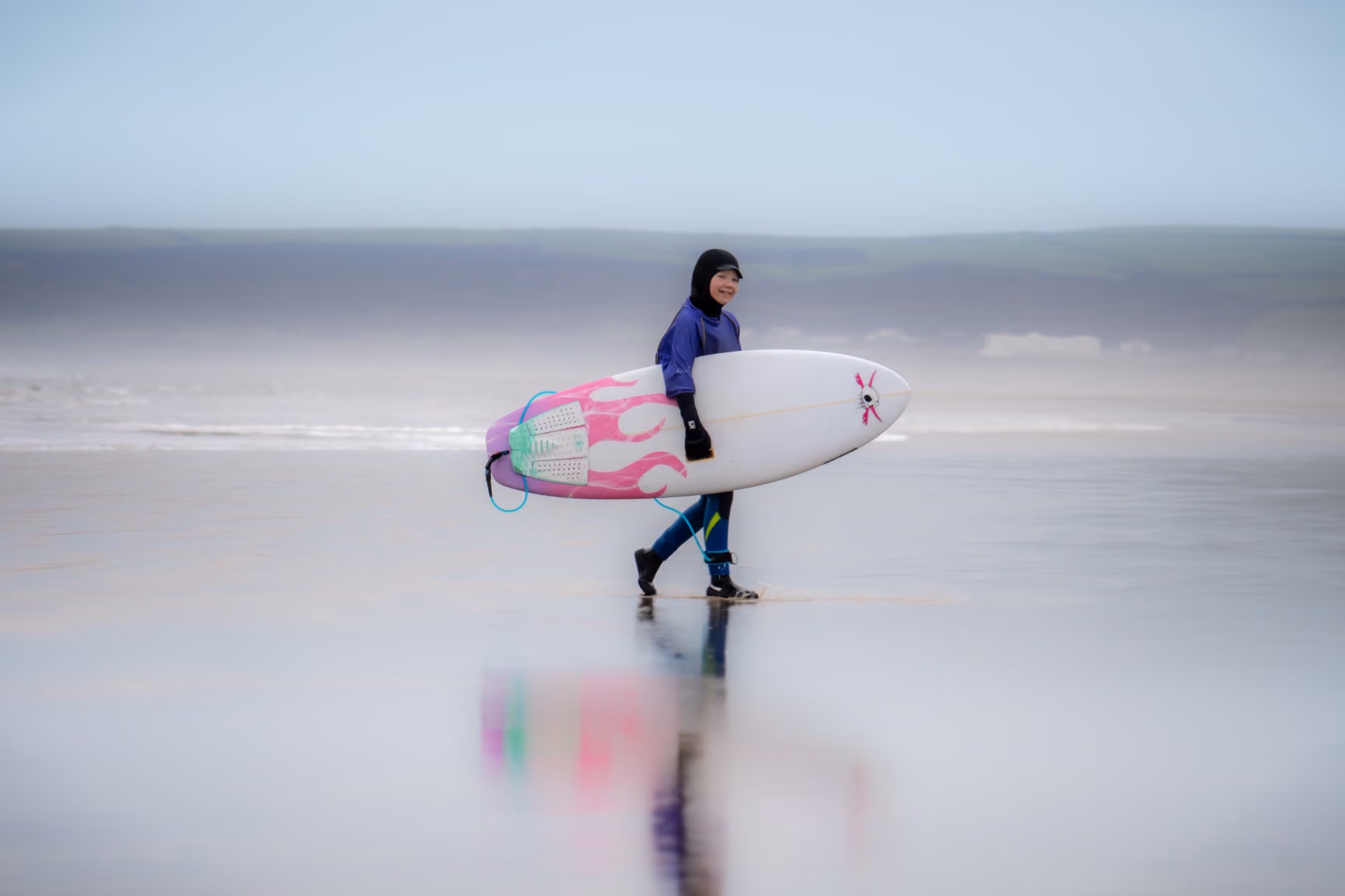 Person wearing a wetsuit and hood walking on wet sand at the beach carrying a white surfboard with pink flame designs and green foot grips.