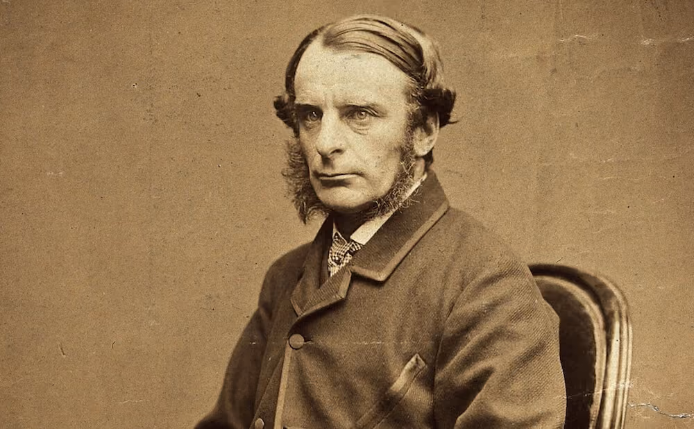Black and white portrait photograph of a Victorian-era man with prominent sideburns, wearing a buttoned coat and seated on a chair, looking slightly to the left.