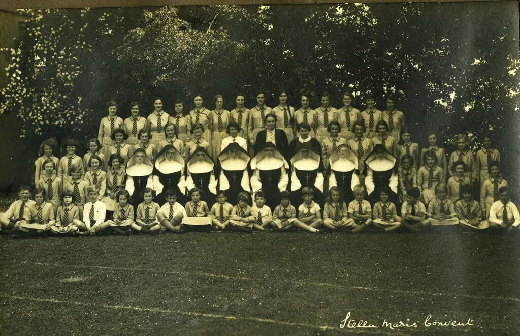 Black and white group photo of a large assembly of schoolgirls and nuns outdoors on grass, surrounded by trees. The girls are dressed in uniform shirts and striped ties. Seven nuns in habits with large white headpieces and crosses sit in the middle row. The inscription at the bottom right reads 'Stella Maris Convent.'