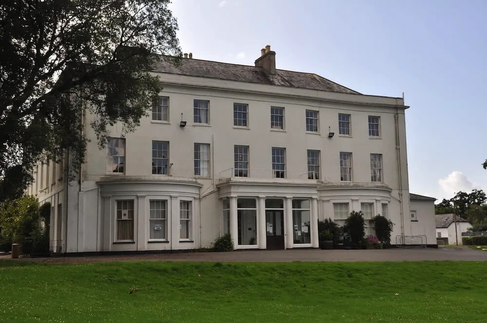 Large three-storey white building with multiple tall windows, a central entrance with columns, surrounded by green lawn and trees under a clear sky.