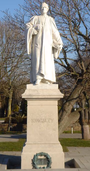 White statue of a man in robes holding a book in one hand and a staff in the other, standing on a pedestal inscribed with 'KINGSLEY', set in an outdoor park with leafless trees in the background.