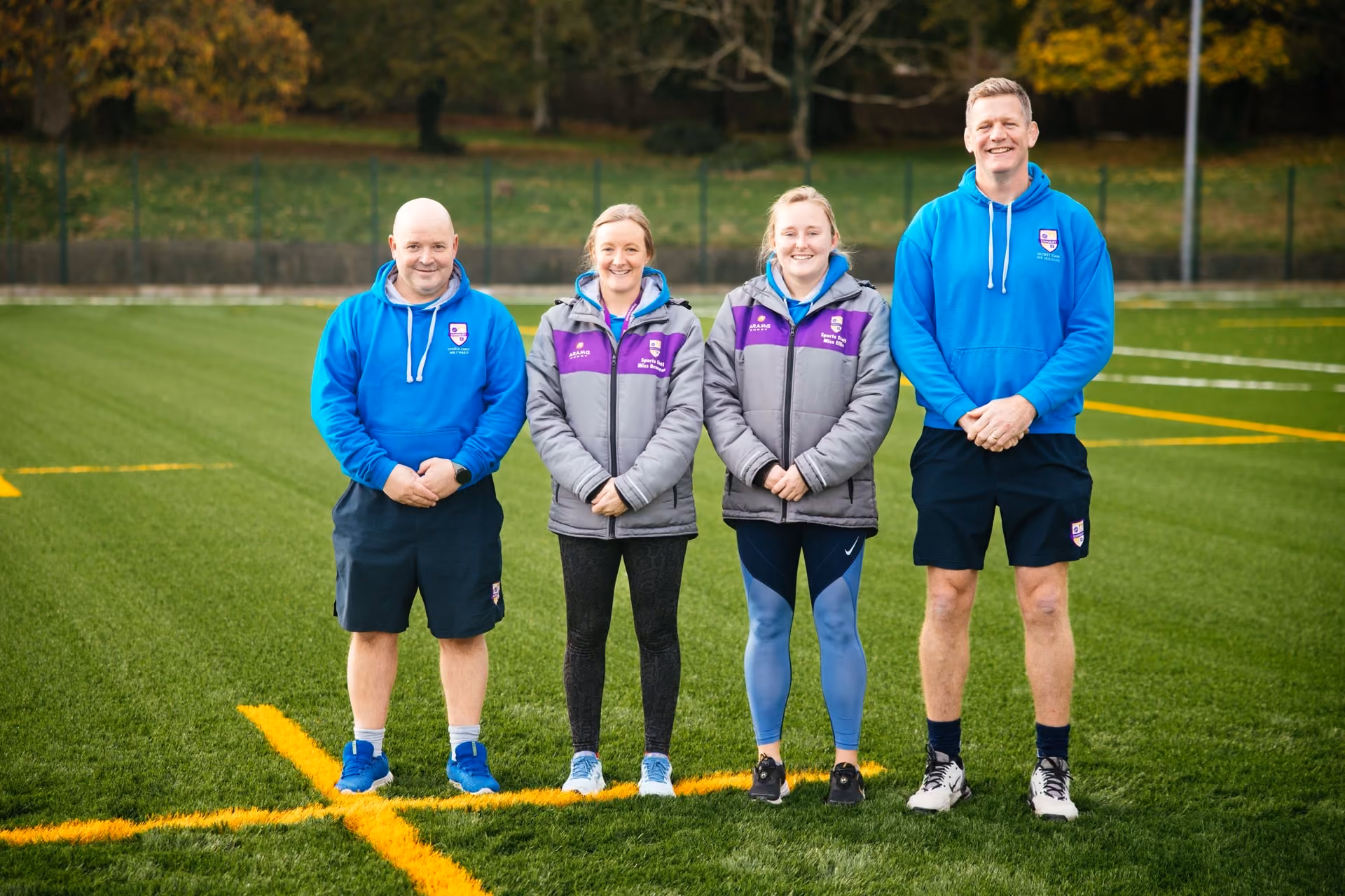Four people standing on a green astro turf sports field, two men wearing blue sports hoodies and shorts, two women in grey jackets with purple accents, all smiling at the camera.
