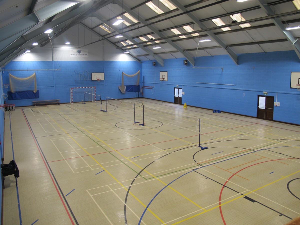 Indoor sports hall with blue walls and ceiling skylights, marked for multiple sports including basketball and badminton, with three basketball hoops and badminton nets set up.