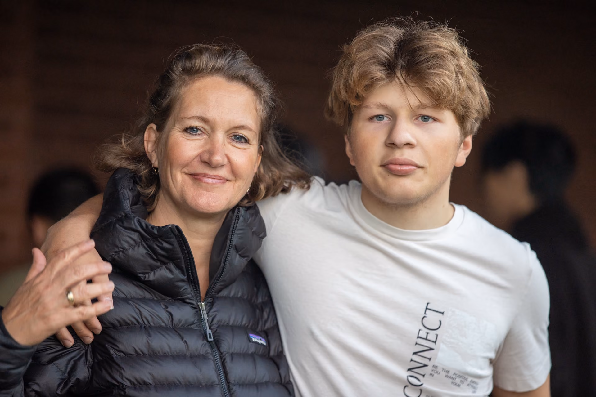 A smiling woman in a black puffer jacket with her arm around a young man in a white T-shirt with the word CONNECT printed vertically, both facing the camera.