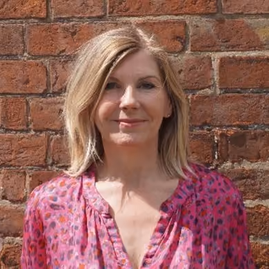 Smiling woman with shoulder-length blonde hair wearing a pink patterned blouse standing in front of a red brick wall.