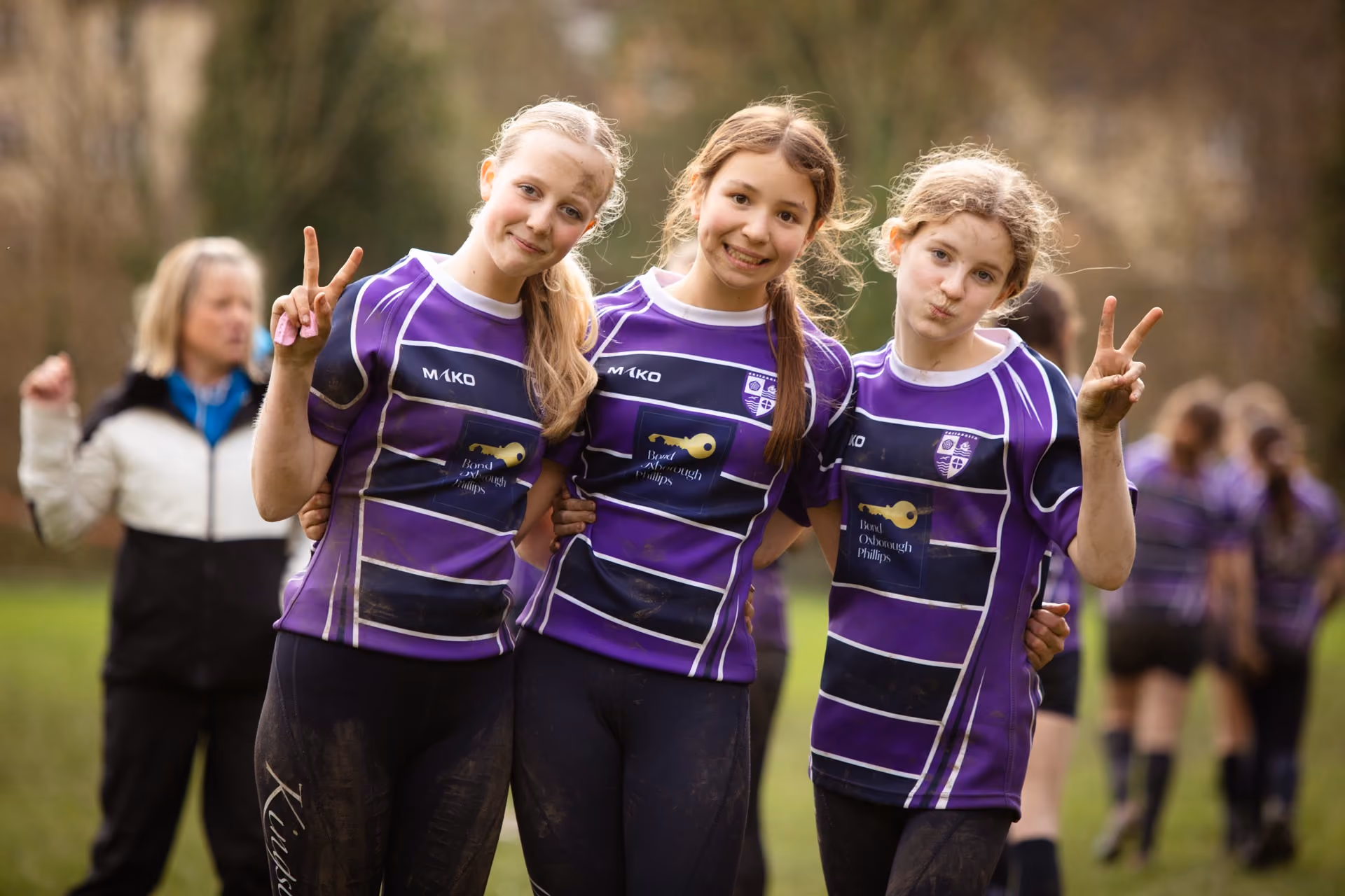 Three young girls in matching purple and black rugby uniforms pose outside on a grassy field, smiling and making peace signs with their hands.