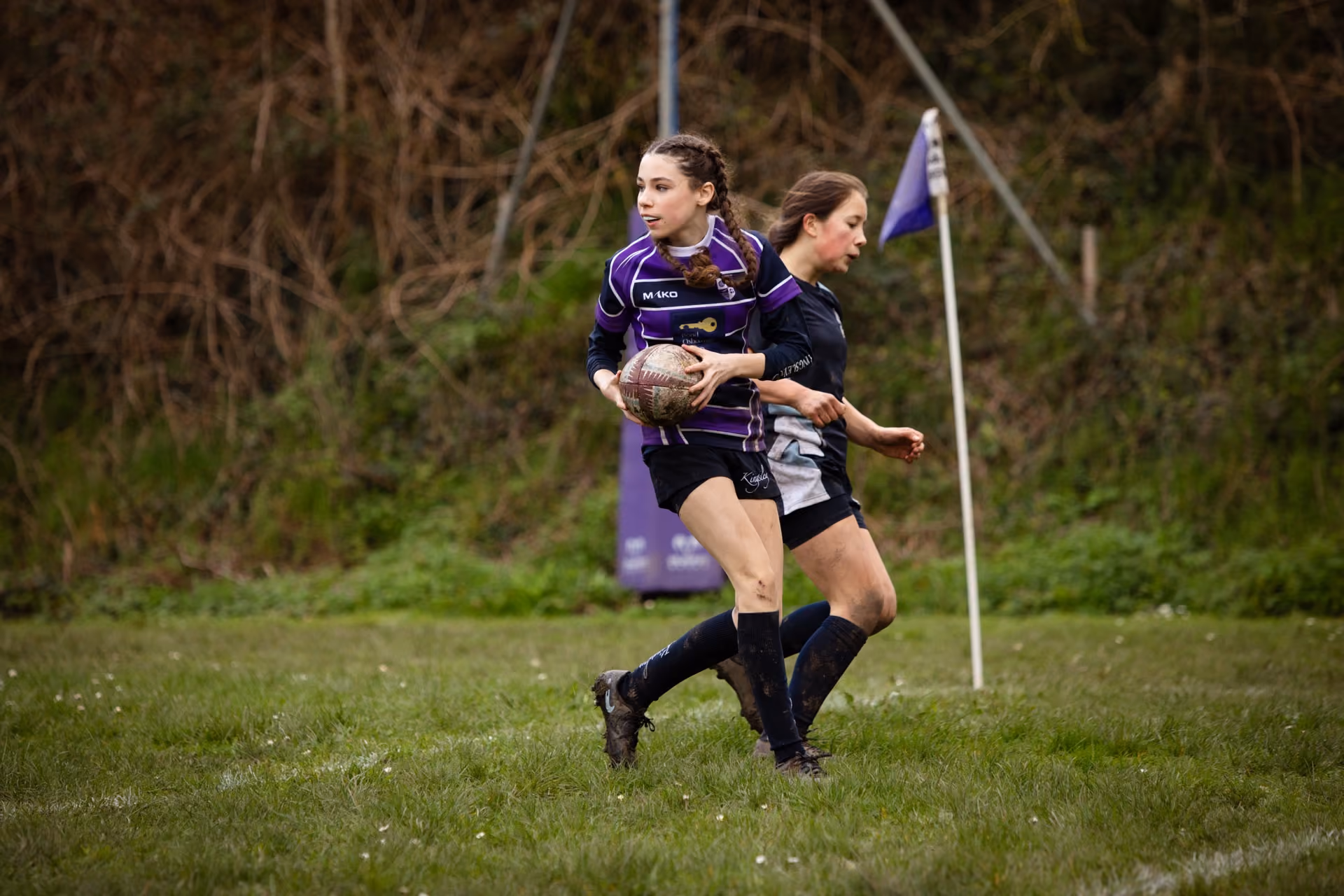 Two female rugby players on a grassy field near a corner flag, one in purple and black holding the ball and the other in black closely defending.