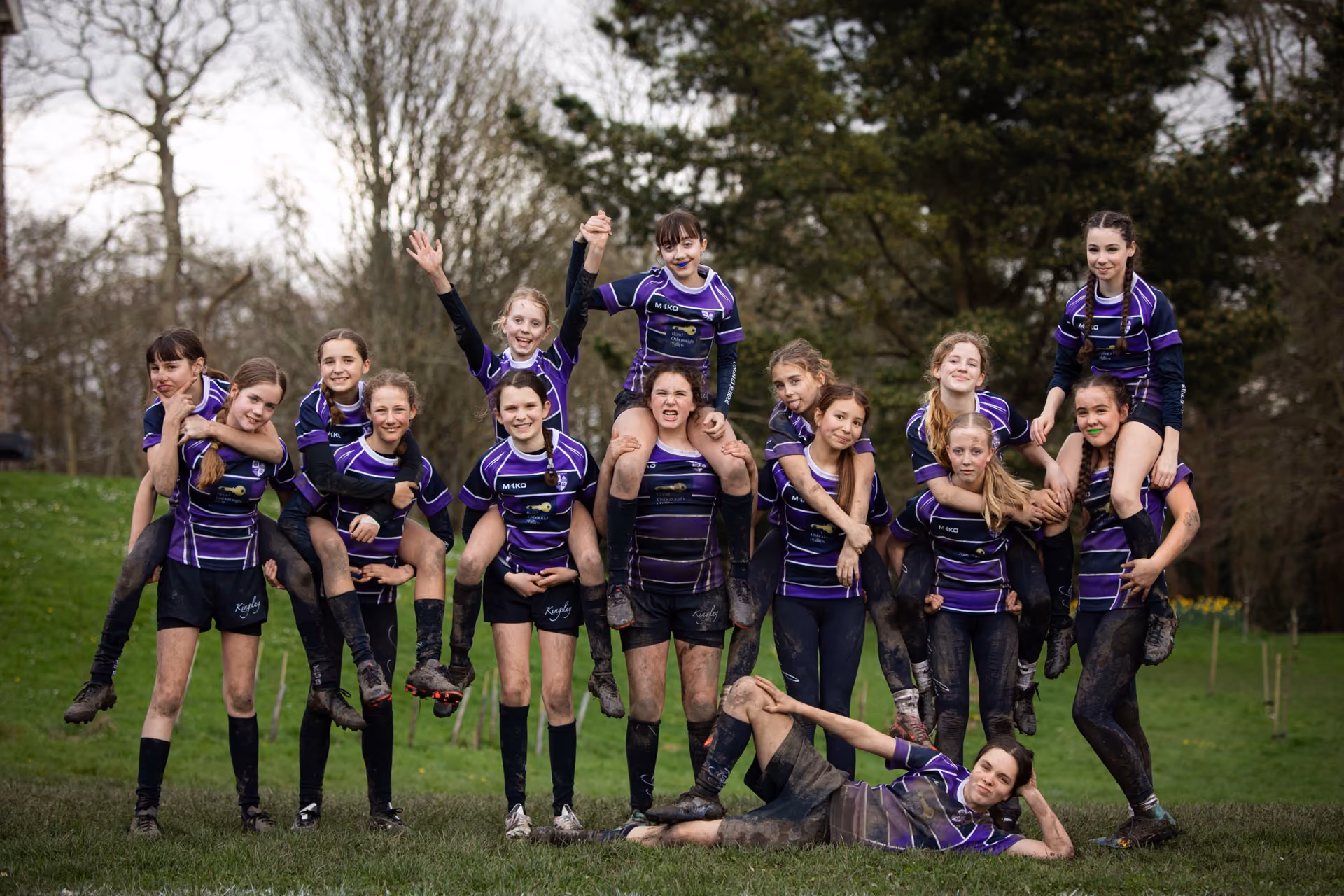 Group of young female rugby players in purple and black uniforms posing outdoors on grass with some teammates piggybacking others and one lying on the ground, all smiling.