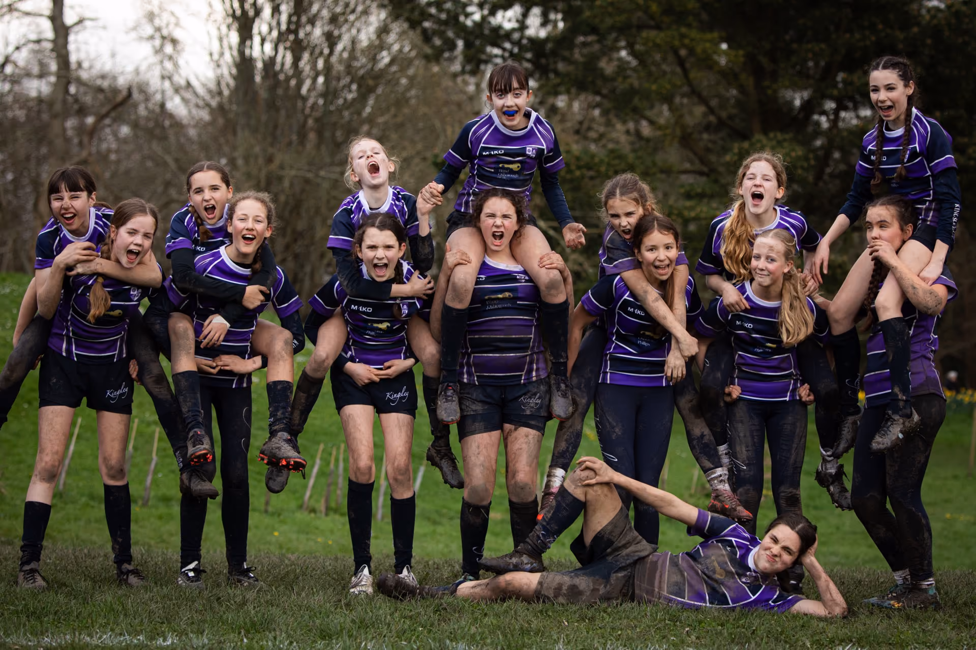 A group of young female rugby players in purple and black uniforms, some carrying teammates on their backs, posing and cheering on a muddy grass field with trees in the background.