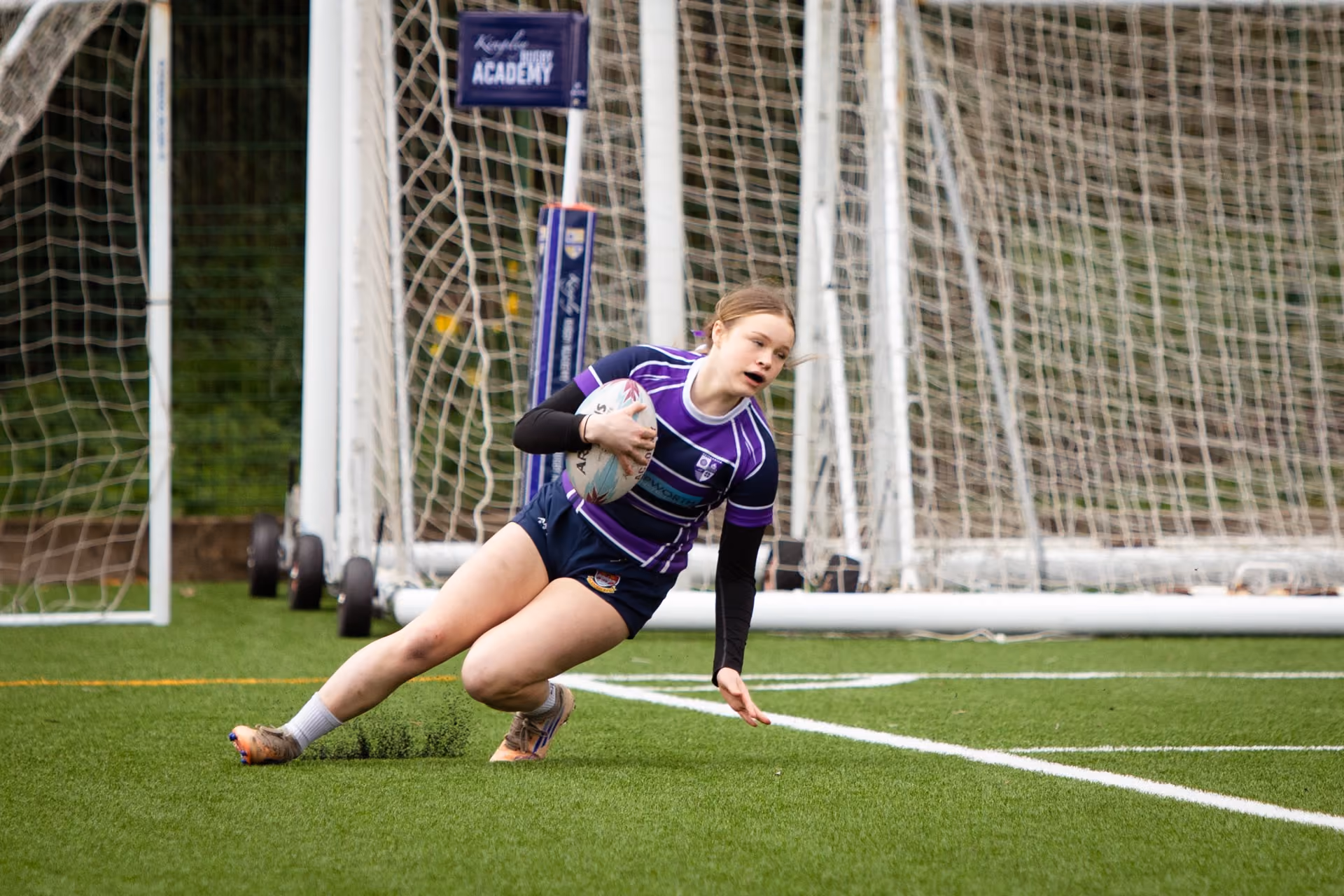 Female rugby player in a purple and black striped jersey lunging with a rugby ball on a green artificial turf field near white goalposts and a net.