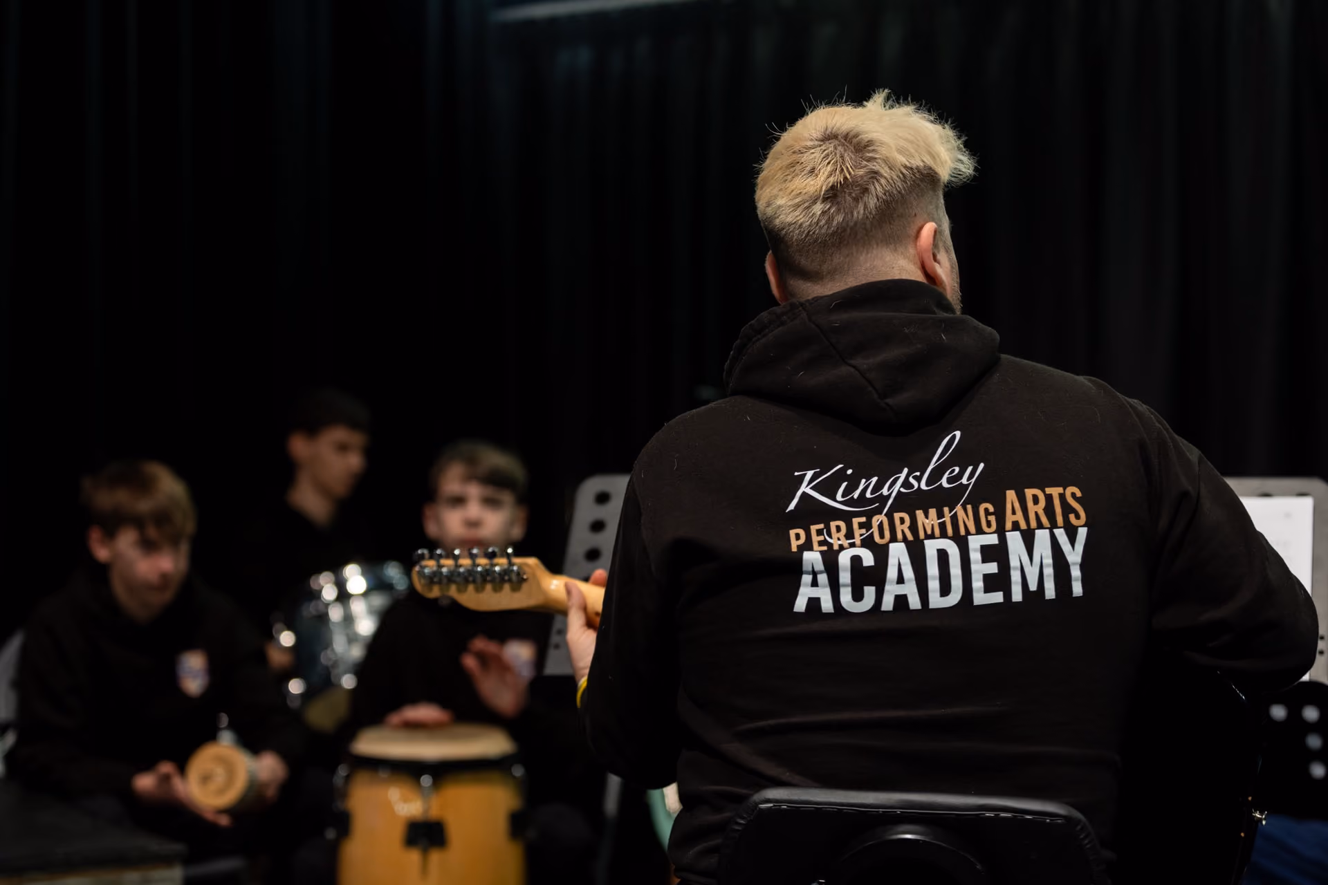Person with blonde hair wearing a Kingsley Performing Arts Academy hoodie playing an electric guitar facing a group of boys playing percussion instruments in a music rehearsal room with black curtains.