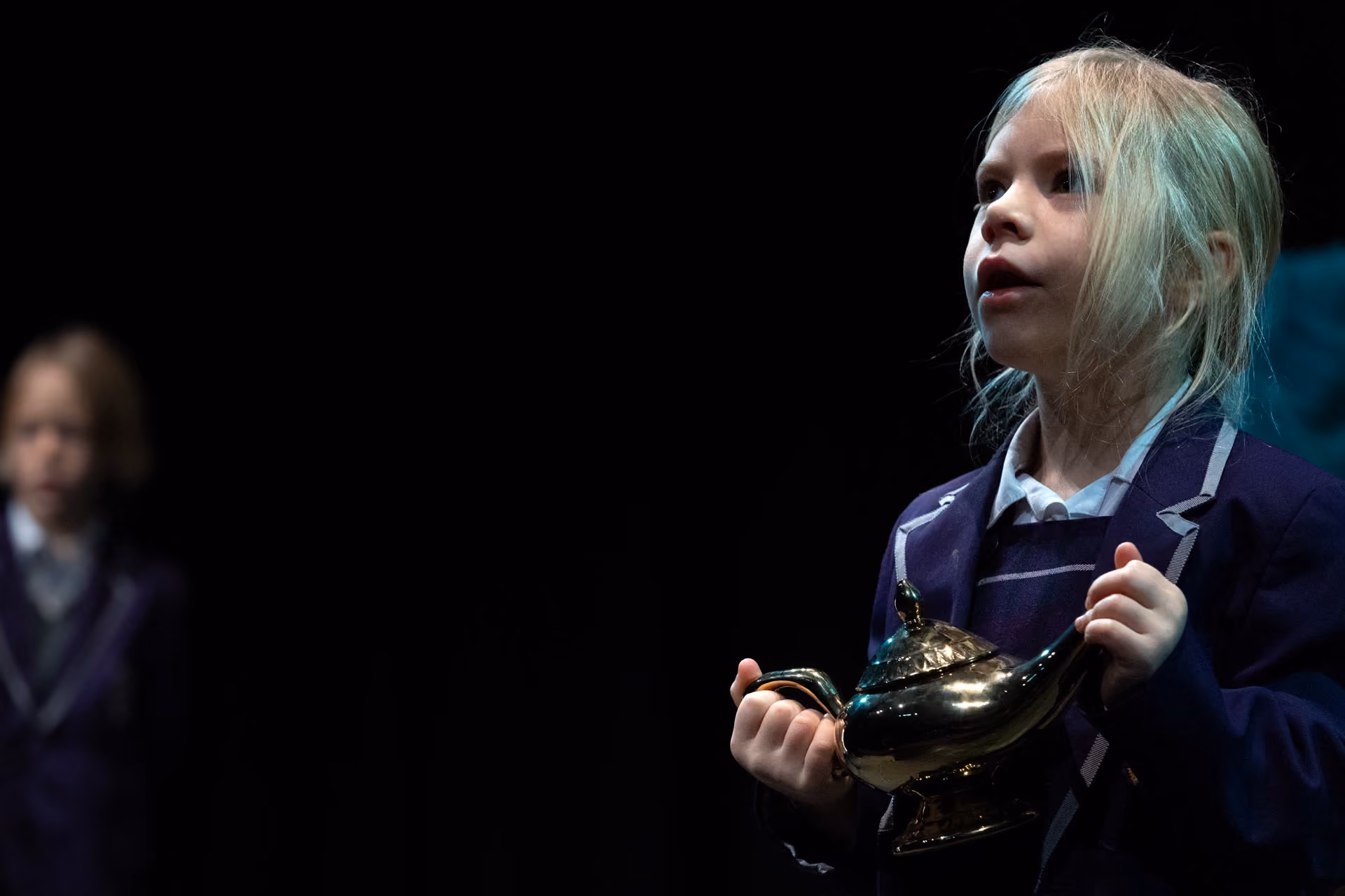 Young girl in school uniform holding a shiny brass genie lamp on stage with a black background, appearing to perform theatrically.
