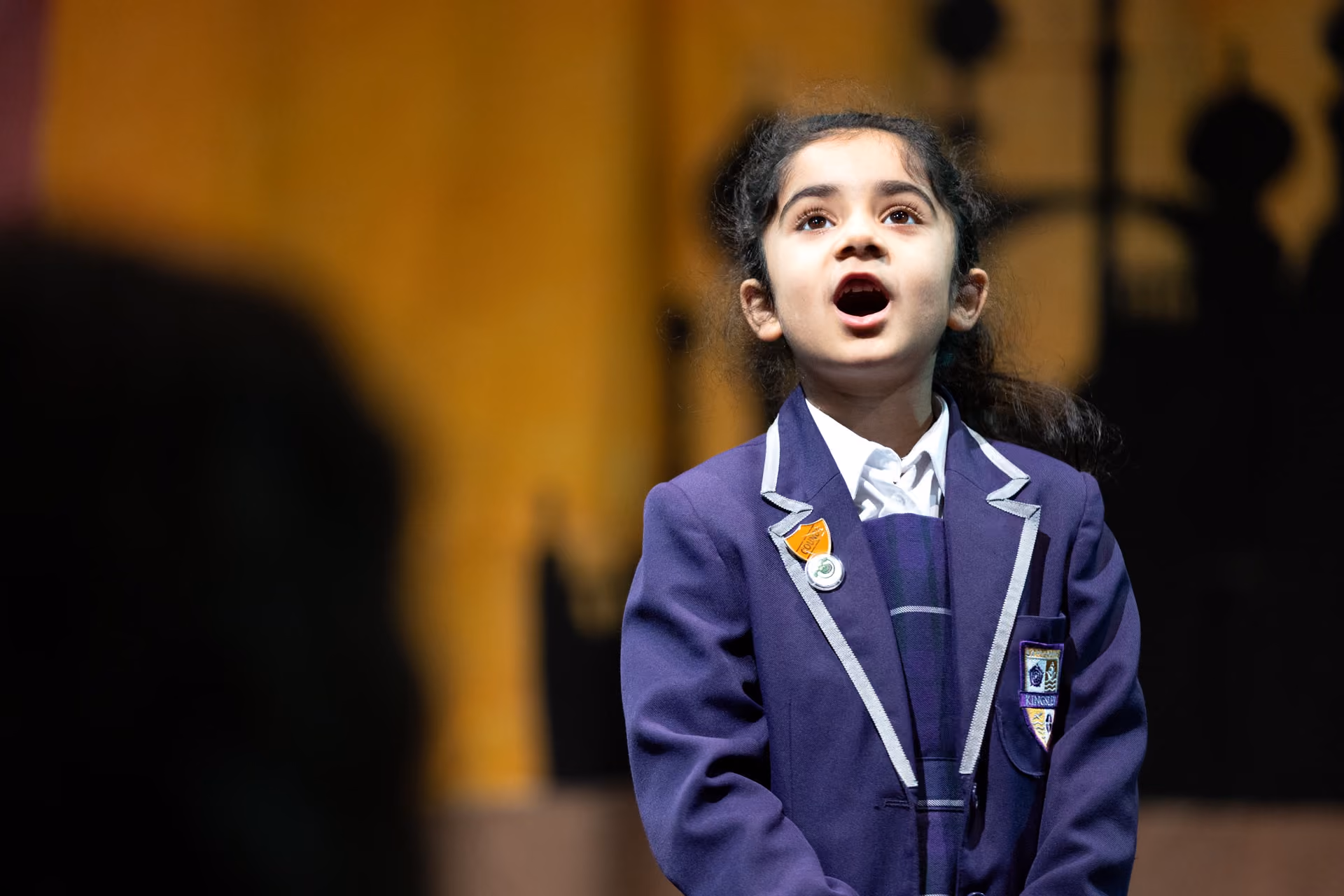 A young girl in a purple school blazer and white shirt singing with her mouth open, against a softly blurred background.