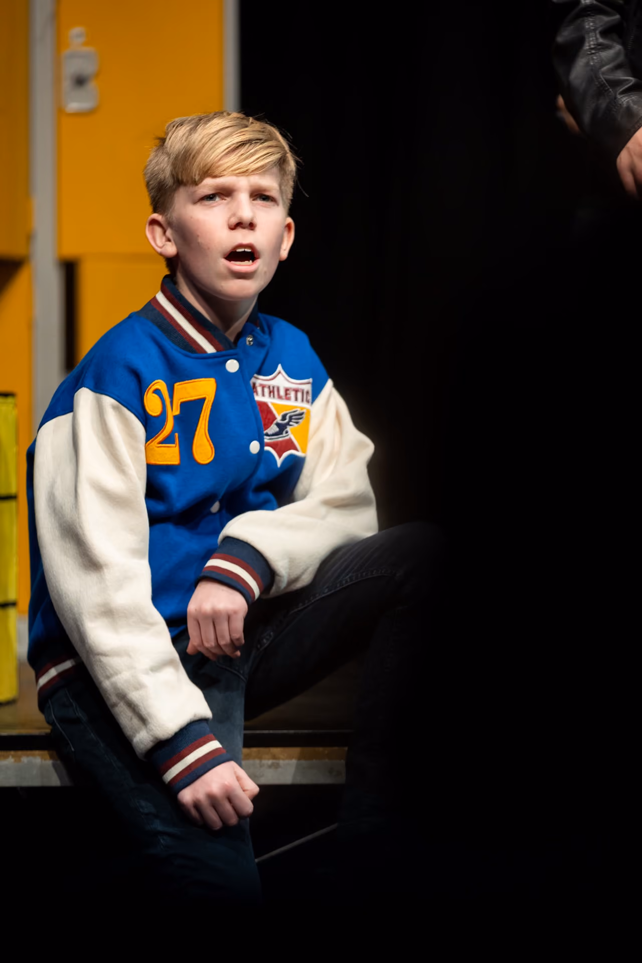 Blond boy wearing a blue and white varsity jacket with number 27 and patches, kneeling and speaking with a serious expression.