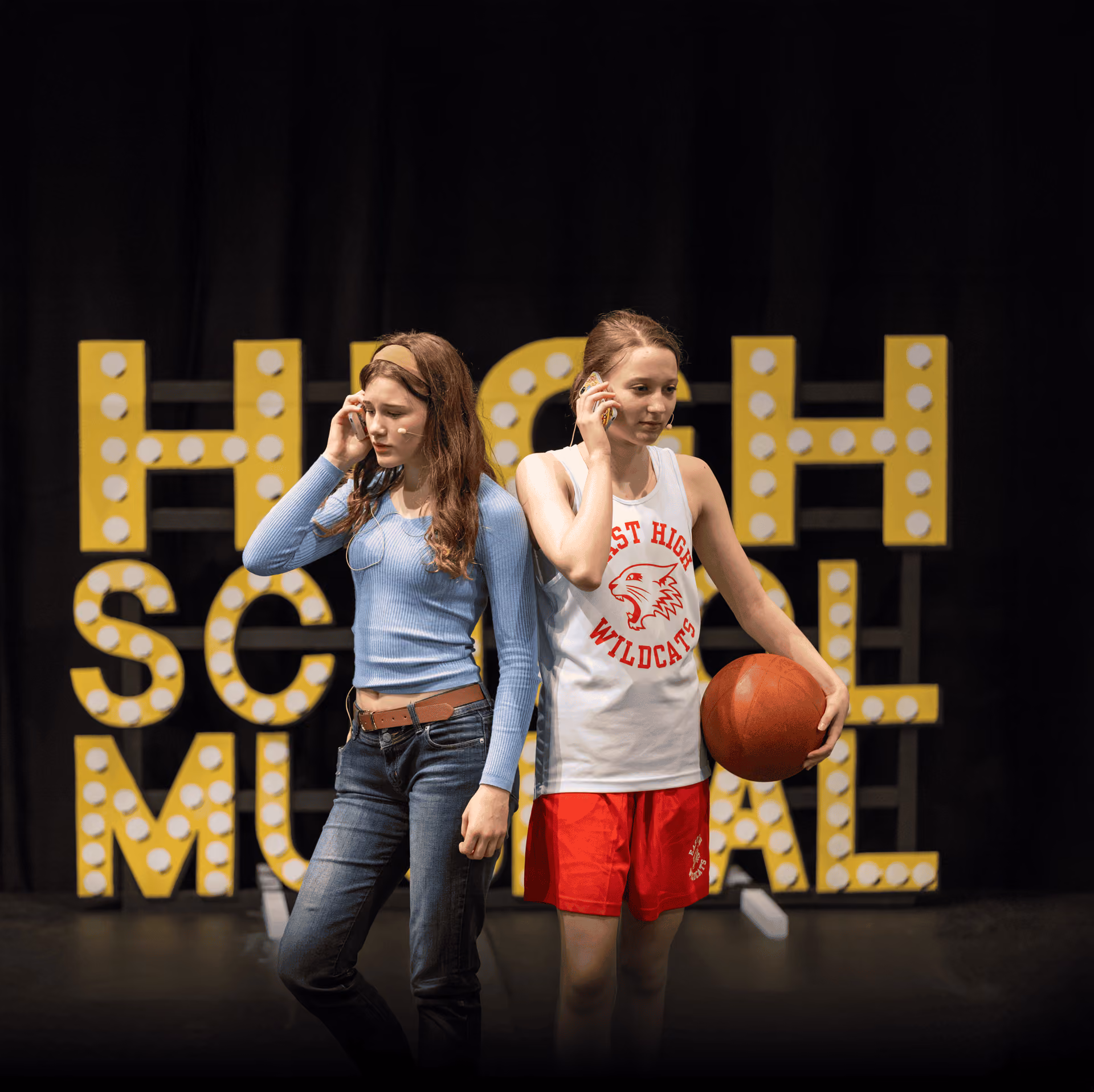 Two teenage girls standing back to back on stage taking phone calls; one wears a blue long-sleeve shirt and jeans, the other wears a white basketball jersey with red shorts holding a basketball, with illuminated letters spelling 'HIGH SCHOOL MUSICAL' behind them.