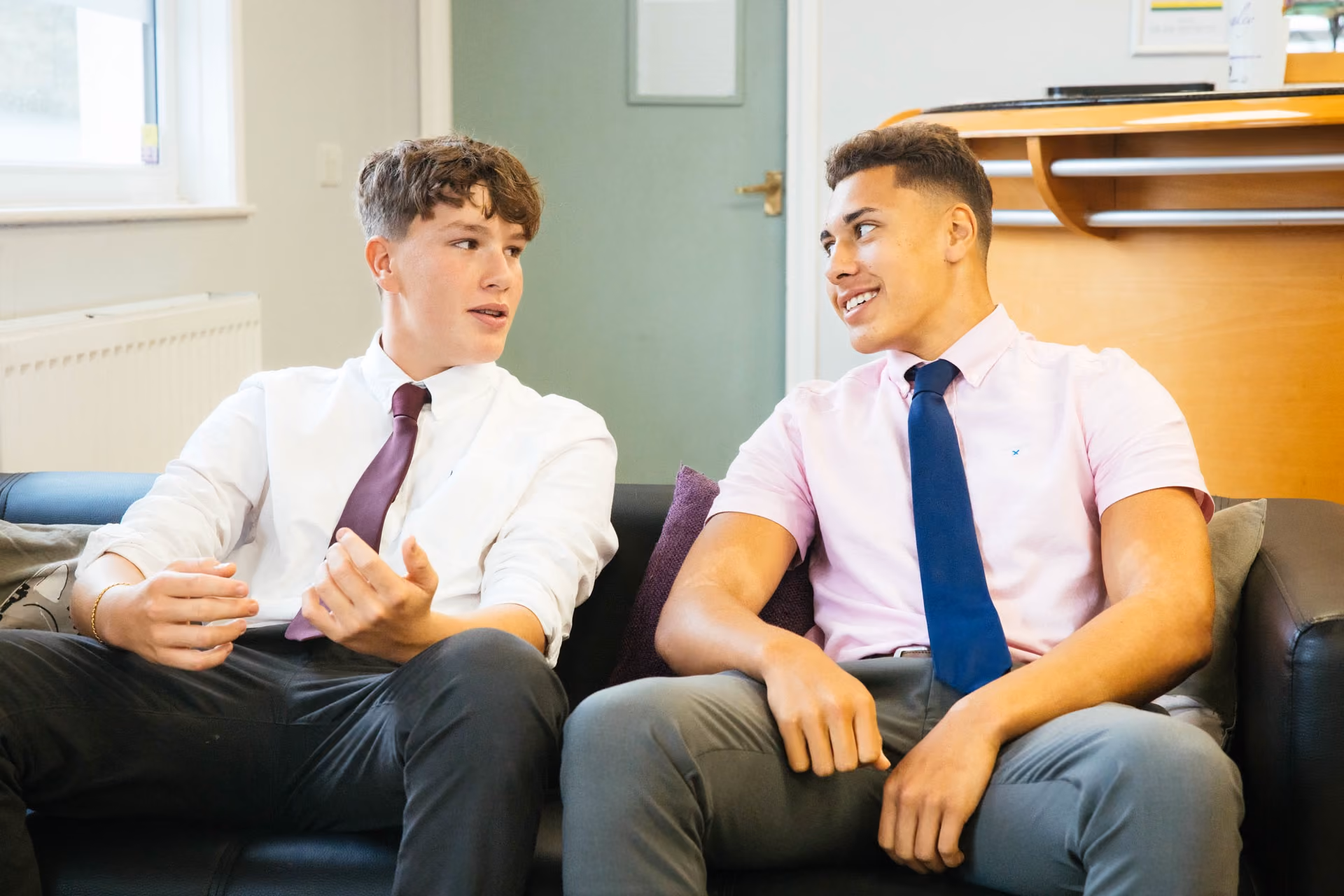 Two young men sitting on a black leather sofa facing each other, engaging in conversation; one wears a white shirt with a maroon tie and the other a light pink shirt with a blue tie.
