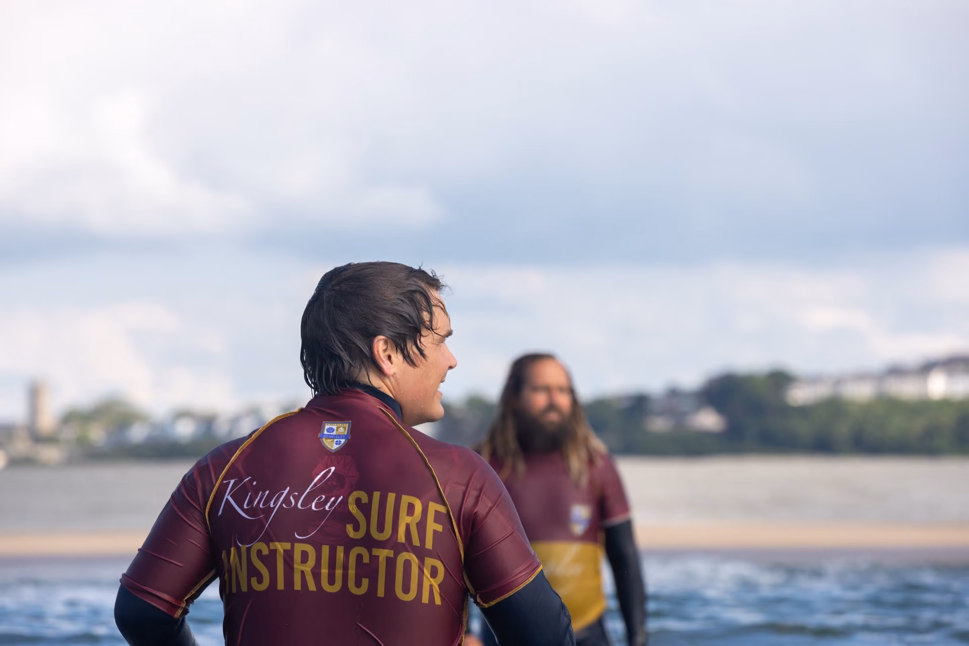Back view of a man wearing a maroon and yellow wetsuit with the words 'Kingsley Surf Instructor' on the back, standing near the water with another man blurred in the background.