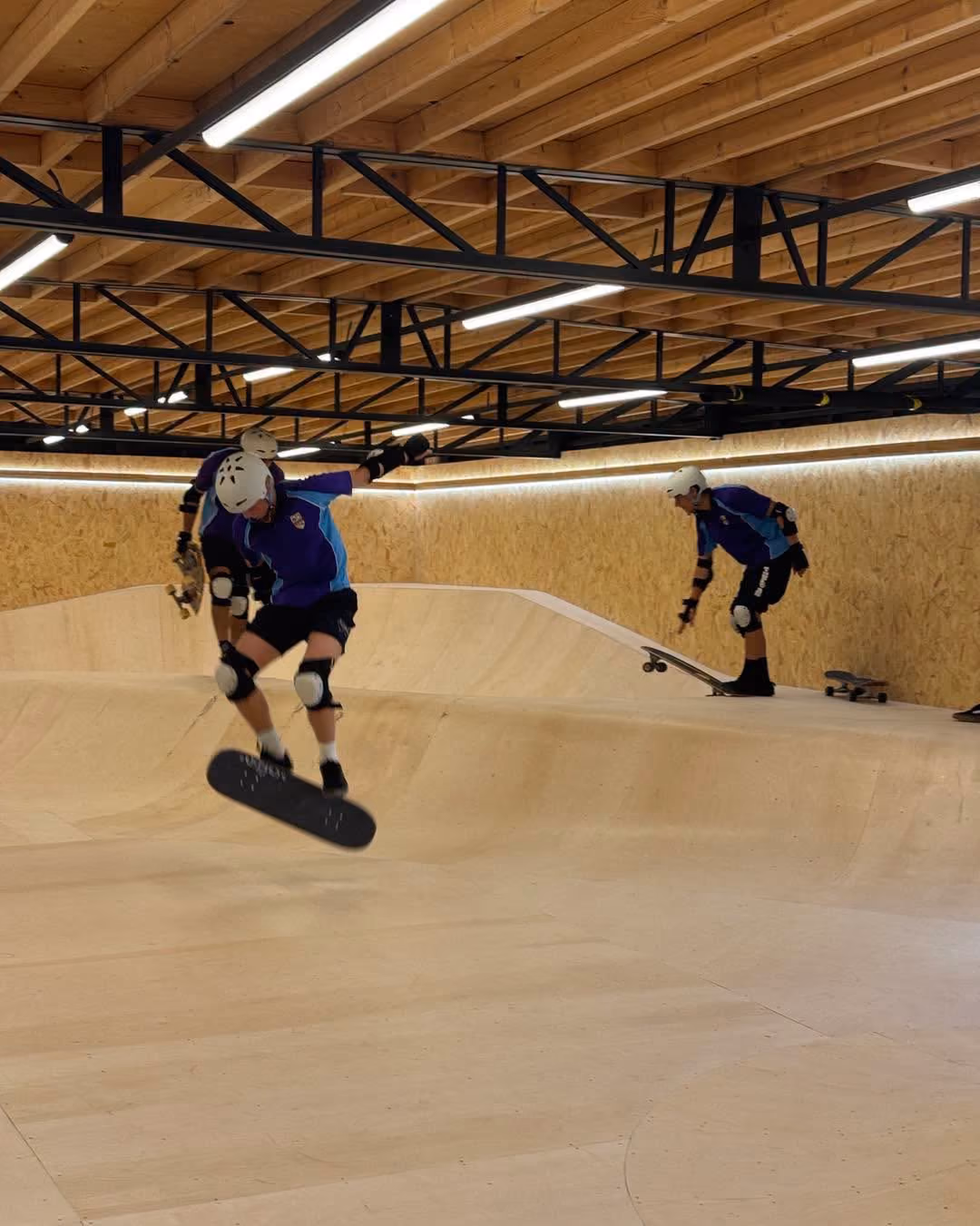Indoor skatepark with three skateboarders wearing helmets and protective gear, one mid-air performing a trick, another preparing to skateboard on a wooden ramp, and a third person in the background.