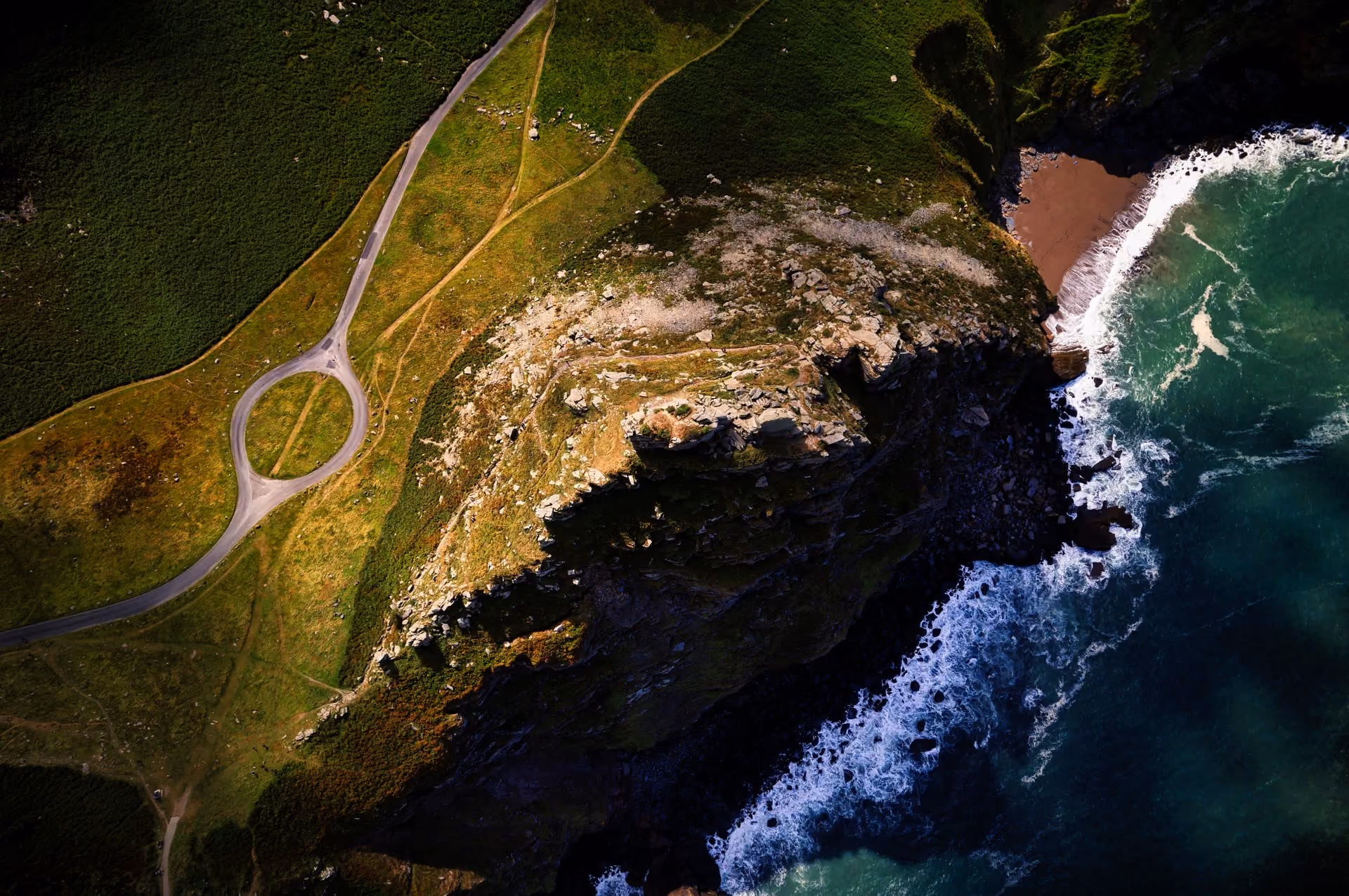 Aerial view of a coastal cliff with green grass and rocky terrain beside ocean waves crashing on the shore, featuring a roundabout road near the cliff edge.