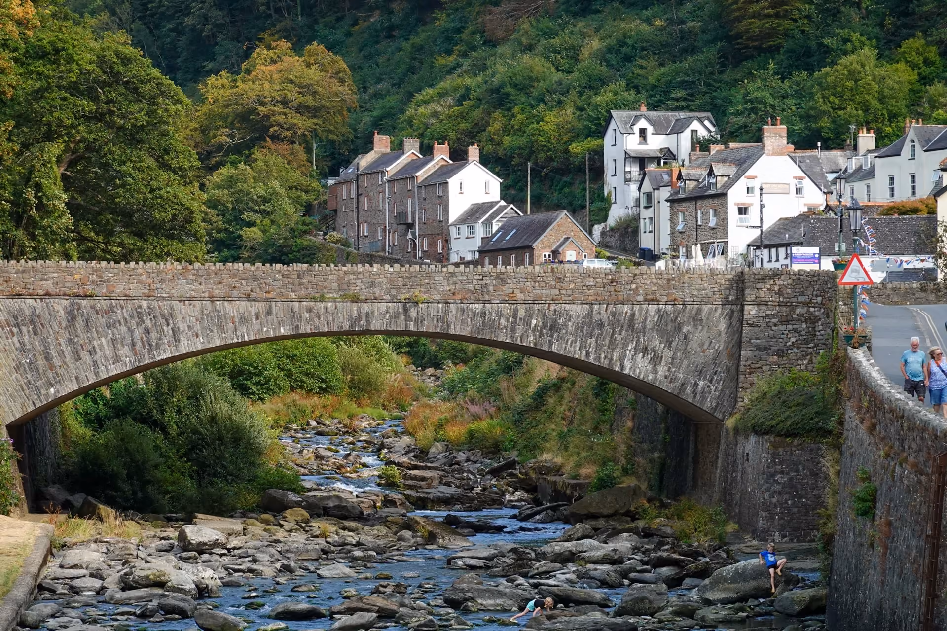Stone arch bridge crossing a rocky river with green foliage and trees alongside, and a quaint village with white and brown houses in the background under a cloudy sky.