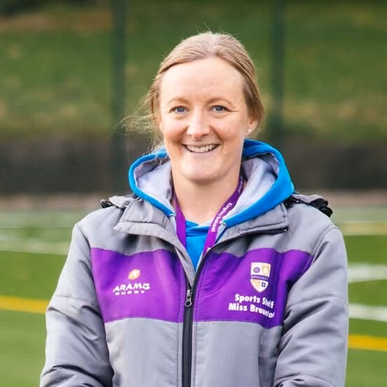 Smiling woman wearing a grey and purple jacket with a sports staff badge, standing on a sports field.
