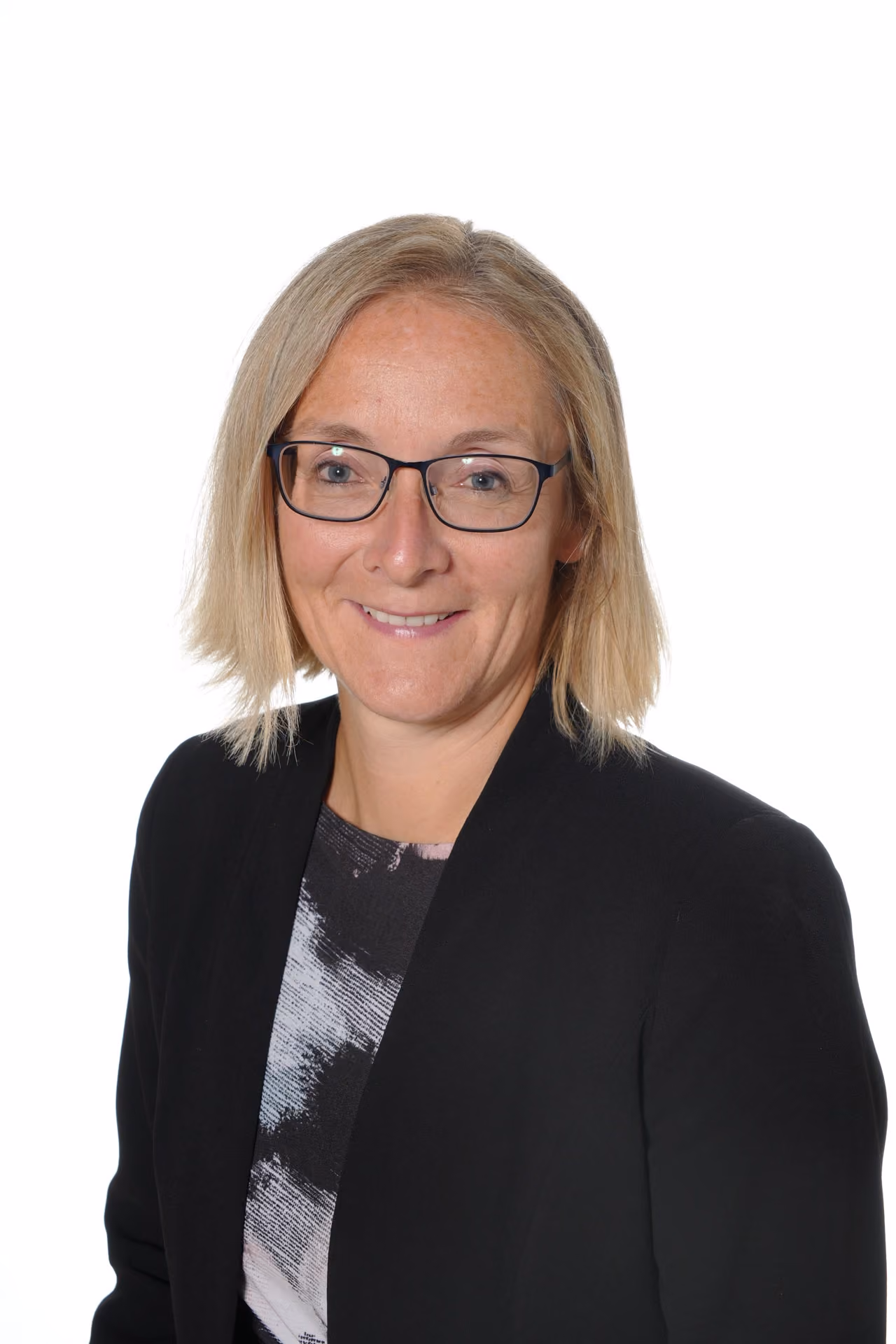 Smiling woman with shoulder-length blonde hair wearing glasses, a black blazer, and a patterned top against a white background.