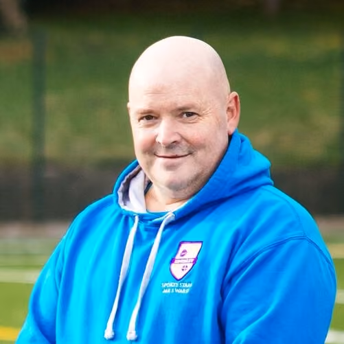 Smiling bald man wearing a bright blue hoodie with a sports team logo on the chest, standing outdoors with a blurred green background.