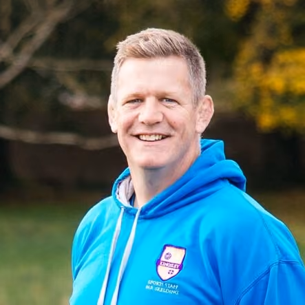 Smiling man with short blond hair wearing a bright blue hoodie standing outdoors with blurred green and yellow foliage in the background.