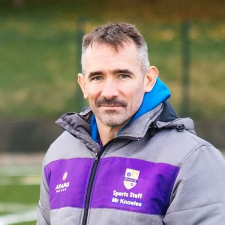 Man with short dark hair and moustache wearing a grey and purple sports jacket with hood, standing outdoors in a sports field setting.