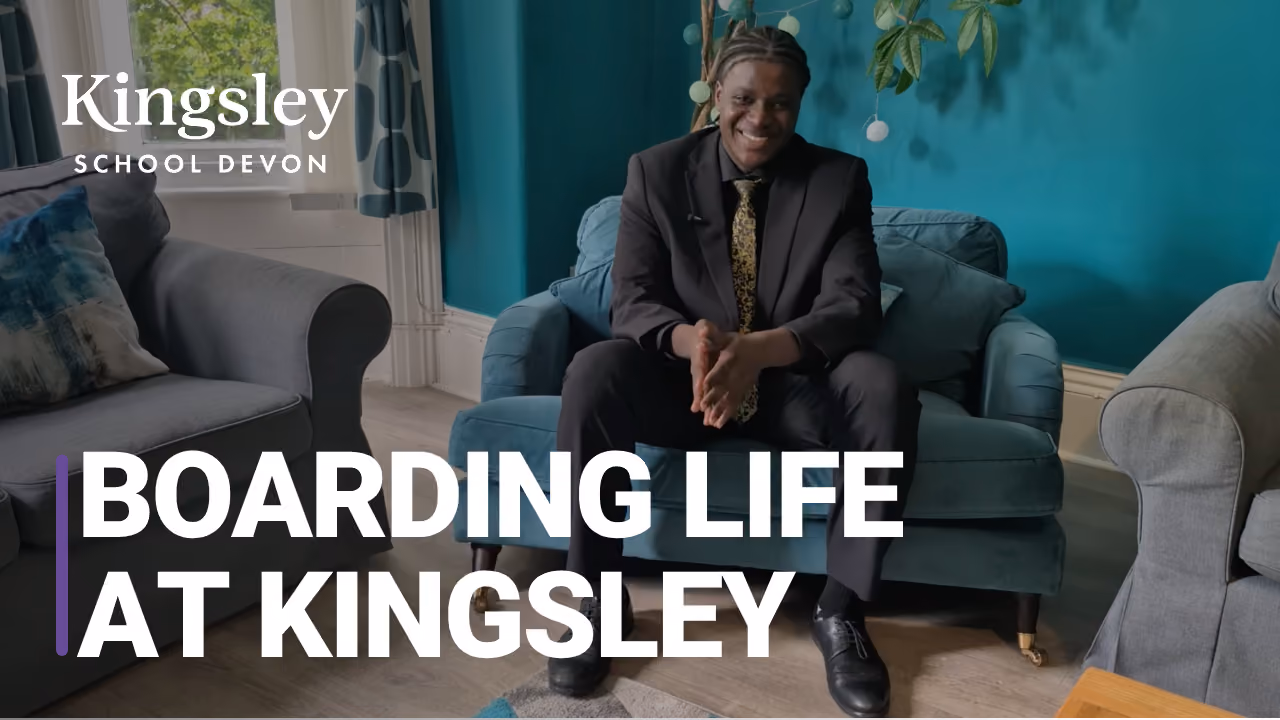 A young man in a dark suit and patterned tie sitting on a teal sofa smiling, surrounded by armchairs and a plant in a room with teal walls and wooden flooring, with text 'Kingsley School Devon' and 'Boarding life at Kingsley'.