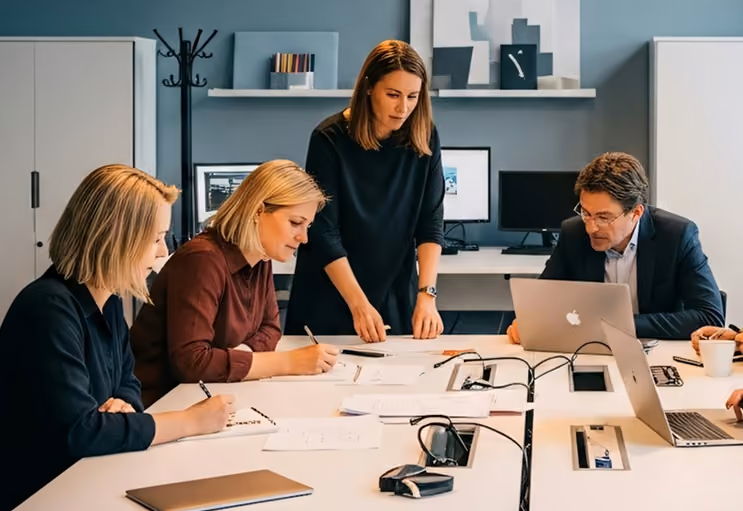 Four professionals collaborating at a table with laptops and papers in an office meeting room.