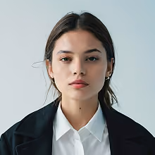 Portrait of a young woman with dark hair pulled back, wearing a white collared shirt and black jacket, looking directly at the camera with a neutral expression against a plain light background.