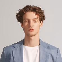 Young man with curly brown hair wearing a light blue blazer over a white t-shirt, posing against a plain light background.