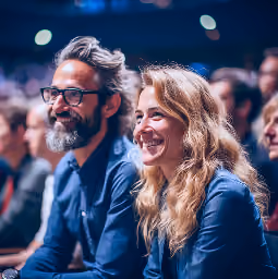 Smiling man with glasses and beard and a woman with long blonde hair sitting together in an audience, slightly out of focus background with other people.