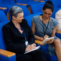 Two professional women sitting in blue auditorium seats, one with gray hair writing in a notebook and the other with dark hair holding a tablet, engaged in discussion.