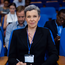 Woman with short gray hair wearing a black blazer and a conference badge sits in a blue chair in an auditorium with other attendees behind her.