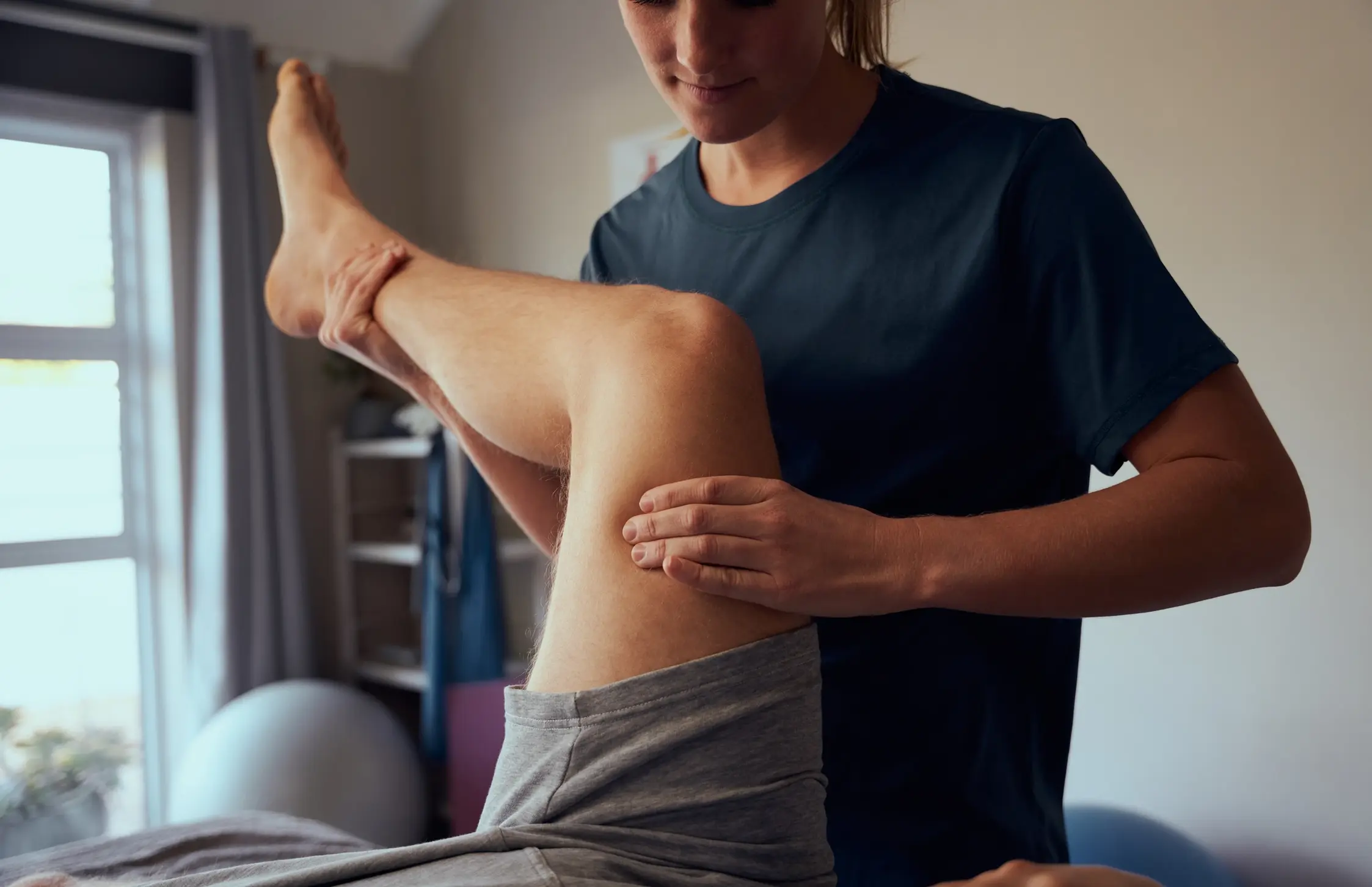 Therapist in navy shirt gently adjusting a patient's bent knee in a treatment room.