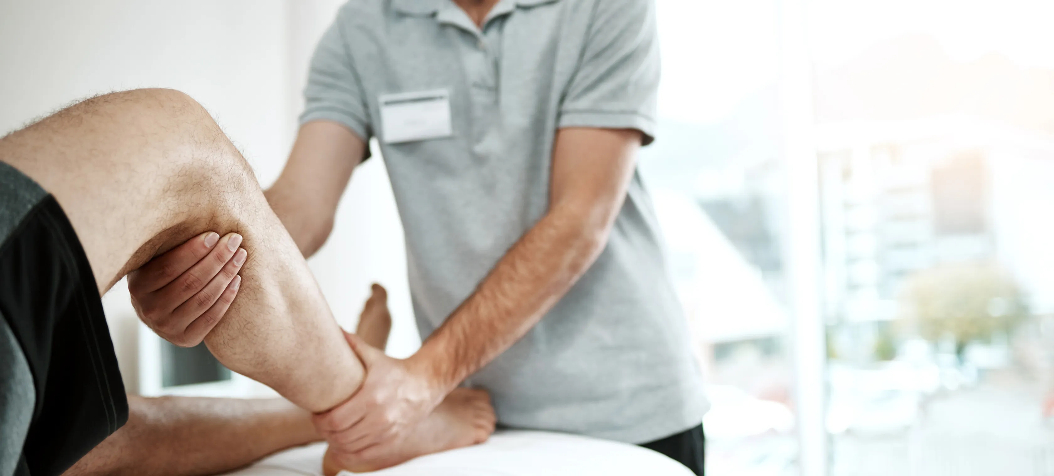 Therapist wearing gray shirt holding patient's bent leg during physical therapy session indoors.