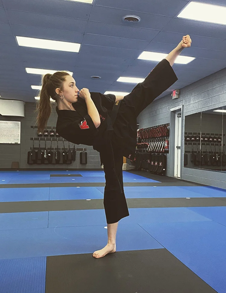 A martial artist performs a high kick while training in a dojo, wearing black clothing on a blue mat with mirrors and storage lockers visible in the background.