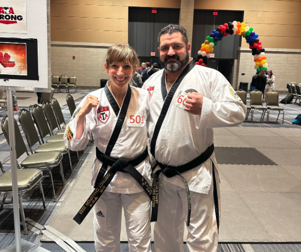 A martial arts instructor and young student wearing white gis proudly hold up their competition medals inside an event venue decorated with a rainbow balloon archway.